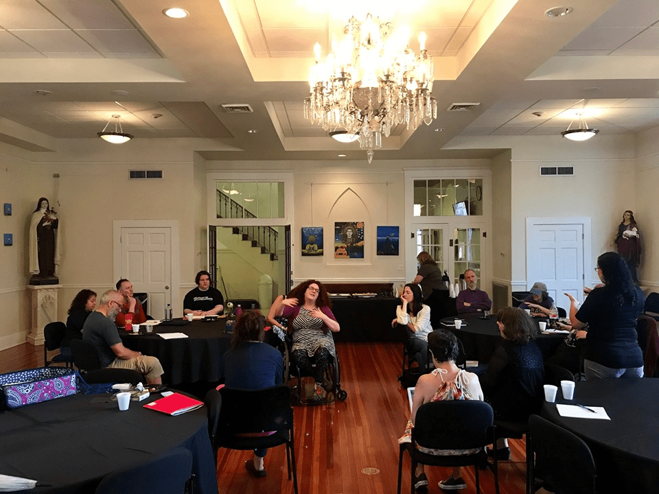 Several poets are standing, sitting, or leaning in black chairs. Jessica is sitting in her wheelchair in the center of the frame, with her head back and her hands actively conveying information.