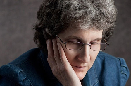 Image of author Kathi Wolfe taken by Alexander Vasiljev. Wolfe's eyes are directed downward. She is wearing glasses and a blue denim top. She has white skin and greying hair. She appears thoughtful.