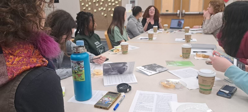 Several workshop participants are clustered around a table in a library. On the table in front of them are many compostable coffee cups, pages of medical records, and various zines.