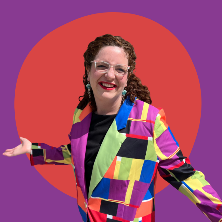 Photo of a white femme person with red hair wearing a very vibrant blazer and octopus earrings